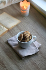 Cup of chocolate chip cookies, open book and reading glasses on the table. Lit candle and bokeh lights in the background. Hygge at home. Selective focus.