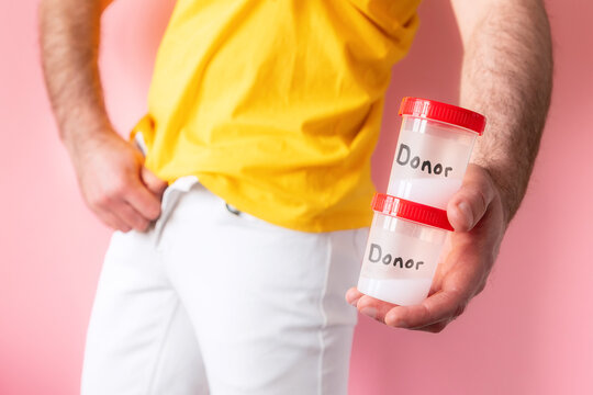 A man in white jeans zip up his fly and holds two plastic jars with sperm samples with text donor in his hands. Pink background. The concept of artificial insemination and sperm donation