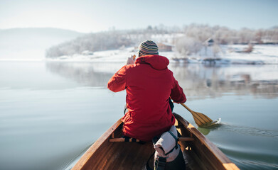 Rear view of man in red jacket paddling canoe on lake winter ride - obrazy, fototapety, plakaty