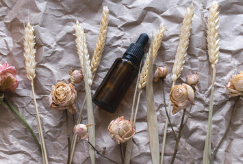 Serum in a brown glass bottle next to spikelets and dry roses on a crumpled beige background. The concept of environmentally friendly natural cosmetics for skin care. Cosmetology. Care for dry skin. 