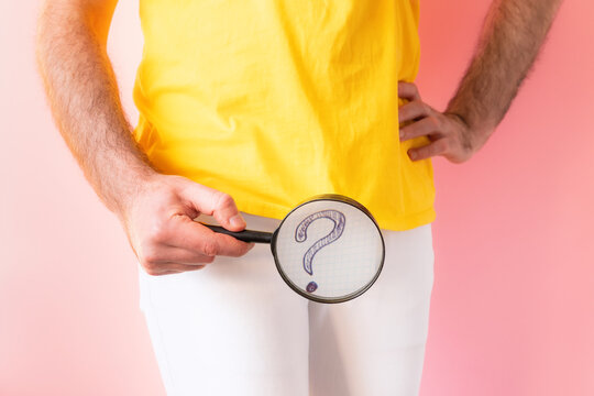 A man in white jeans holds a magnifying glass at the level of the genitals, showing a question mark. Pink background. The concept of impotence and urology