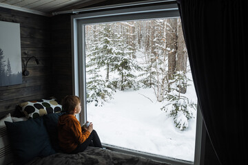 A little dreamy red boy with a mug of cocoa looks through a panoramic window at the forest. Large windows in the house. Winter forest landscape.