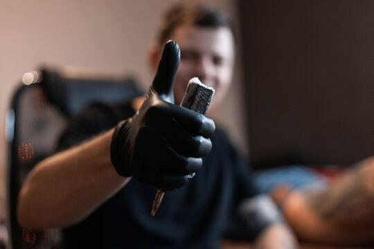 professional tattoo artist man in black gloves holds a tattoo machine and shows thumbs up - like. Working process of creating a tattoo in the studio