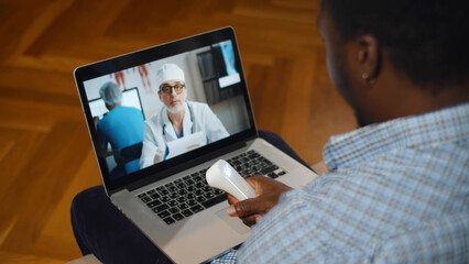 Back view of man measuring temperature making video call with doctor while staying at home. Sick African-American guy with infrared thermometer having online consultation with practitioner