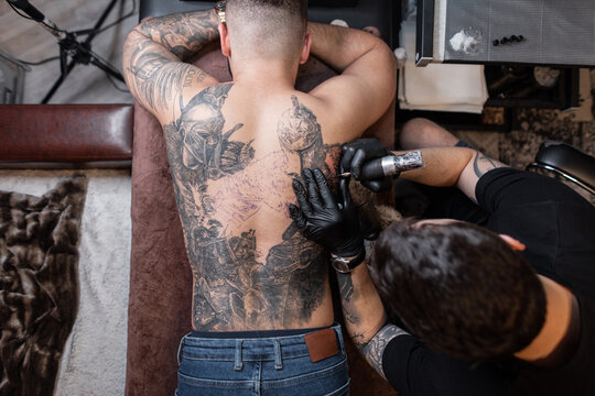 Tattoo artist man with black gloves draws a knight tattoo on the guy's back in the studio, top view. Tattoo artist's workflow