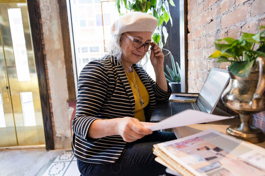 Senior Woman With Paperwork Working At Laptop In Cafe