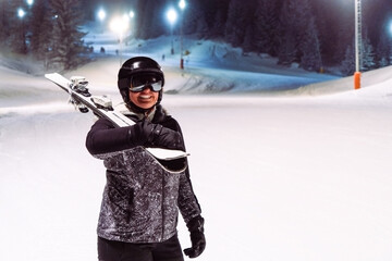 Happy woman skier with skis on her shoulder on a night ski slope - obrazy, fototapety, plakaty