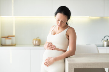 An attractive pregnant woman is standing in the kitchen of her home. She keeps her hand on her stomach so she can feel her baby move. Last months of pregnancy.