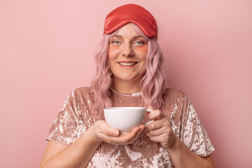 Happy smiling attractive woman wears a sleep mask holding a cup of fragrant coffee or tea in the morning isolated over pink background.