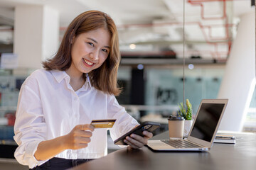 A woman sitting at the counter holding a cell phone and credit card, she is shopping online on a smartphone, she pays for goods and services by credit card. Concept of using credit card for payment.