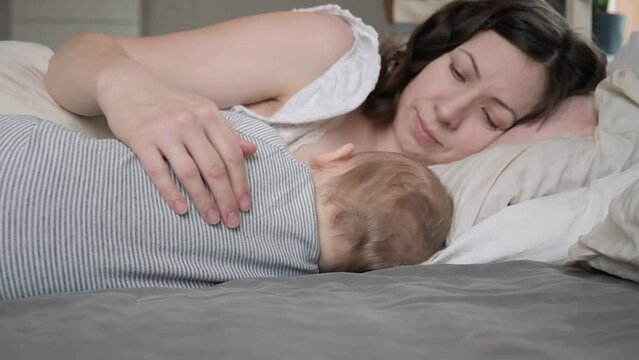 One Caucasian brunette mother in nightwear breastfeeding her tiny newborn, while relaxing on bed. Young woman with baby sleeping at home. Bed-sharing concept. Infant child with his mom in bedroom.