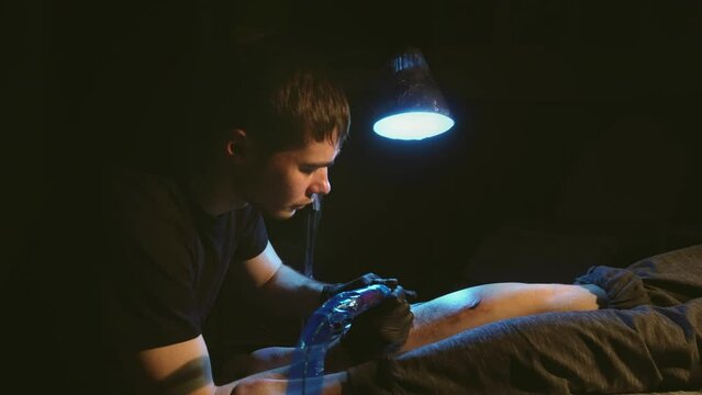 Face portrait of young adult tattoo artist working with rotary machine on customers leg in dark lit room. Master tattooist in gloves with plastic film covered gun tattooing clients shin under lamp.