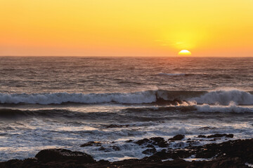 Fototapeta premium A picturesque orange sunset over the Atlantic Ocean, with the sun halfway below the horizon and foaming waves crashing against the dark rocks on the shore