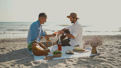 Elegant adult gay couple enjoying romantic date picnic at beach, cheering glasses of wine....