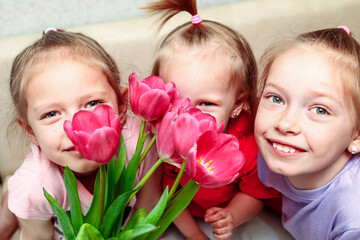 Portraits of two child children and one toddler girl sniffing a large bouquet of pink tulips at home on the sofa. The concept of the eighth of March, a large family and Easter