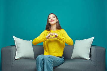 Friendly thankful young girl make fingers heart gesture, showing love, expressing support, gratitude, sitting on sofa