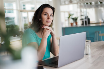thoughtful brooding remote working dark haired woman sitting infront of a laptop or notebook in casual outfit on her work desk in her modern airy bright living room home office with many windows
