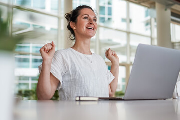 happy joyful successful remote working woman cheerful with fists infront of laptop or notebook in casual outfit sitting on work desk in modern loft living room home office