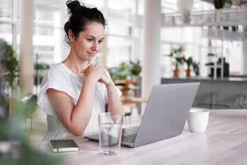 happy smiling remote working woman talking to laptop or notebook in casual outfit sitting on her work desk in her modern loft living room home office having a video chat