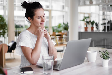 thoughtful brooding remote working dark haired woman sitting infront of a laptop or notebook in casual outfit on her work desk in her modern airy bright living room home office with many windows
