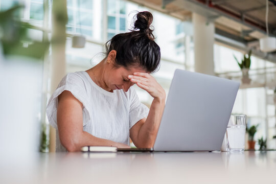 sad depressed remote working woman sitting disappointed with headache infront of a laptop or notebook on her work desk in modern airy bright living room home office with many windows