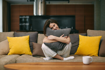 Obraz premium Happy young girl hugging soft pillow sitting on comfortable sofa. Relaxed female resting in modern living room at home