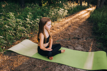 A small, hardworking, beautiful preschool girl, a child athlete sits on a green rug in a lotus position, crossing her legs, performing sports exercises, yoga and gymnastics in the forest at sunset.