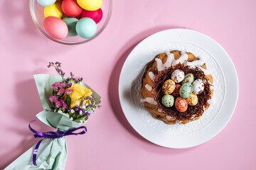 Easter cake with colorful eggs in a chocolate nest on pink background and flowers. Happy Easter concept. Dessert also known as  Kuglof. Top view