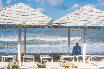 Empty sun loungers under umbrellas on seashore in storm. A lonely man sits with his back and looks...