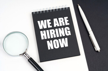 On a black and white surface lies a magnifying glass, a pen and a notepad with the inscription - We Are Hiring Now