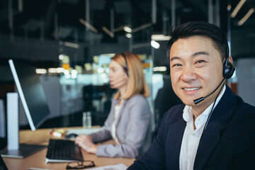Close-up portrait of Asian man in business attire, call center employee looks at camera and smiles, businessman uses computer for video call and headset