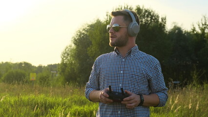 Naklejka na ściany i meble drone pilot wearing headphones and mirrored sunglasses in nature controls drone using remote control. bearded man in plaid shirt flies on drone.