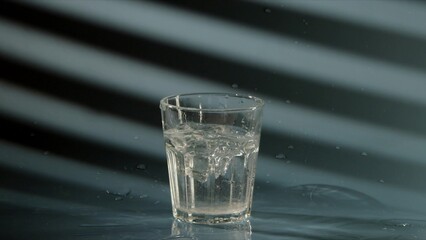 piece of crushed ice falls into transparent glass with clean drinking water, on blue background, sunlight shines through blinds. Slow motion video of cooling drink.