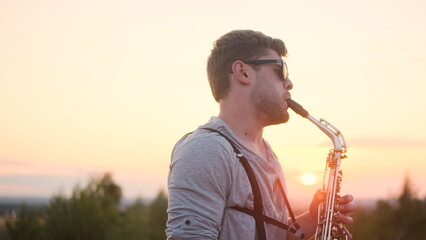 bearded man in glasses and gray turtleneck and suspenders play silver saxophone outdoors in mountains on plain at sunset or sunrise, gentleman play sax with and is satisfied, musical improvisation - obrazy, fototapety, plakaty