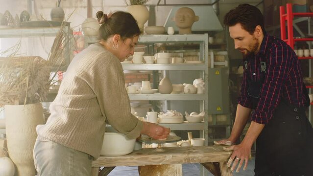 Young woman, potter, is giving masterclass to man, showing how to work on pottery wheel, giving some advice to a beginner, Foreground, Slow motion.