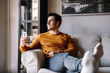 young latin man in sofa using his cellphone and smiling, he is leaning legs on the table with no shoes