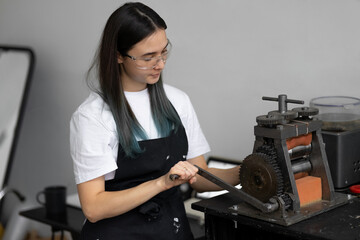 Happy young woman in black work apron, white t-shirt and safety glasses. Rolls silver. Authentic jewelry workshop.