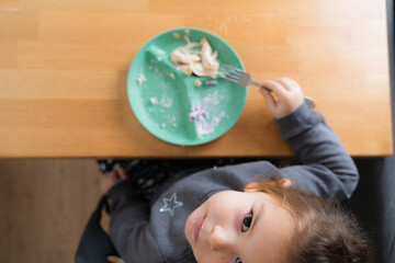 Little child girl sitting at the table and putting spoon at her mouth while eating porridge attentively and with appetite. Stock photo