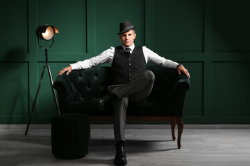 Gentleman in formal clothes and stylish hat sitting on sofa in dark room