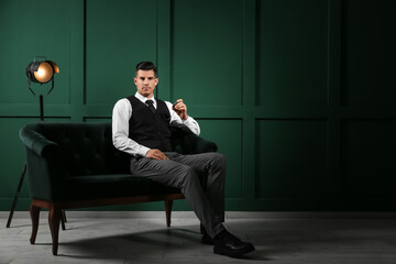 Fashionable gentleman sitting on sofa and holding watch in dark room