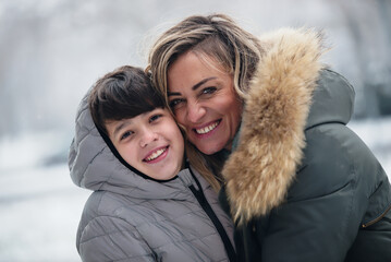Young mom and her teenage son enjoying cold winter weather in the park, hugging and smiling