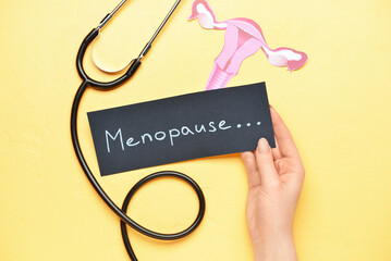 Woman holding paper with word MENOPAUSE, stethoscope and uterus on yellow background
