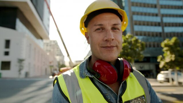 Male worker standing at construction site on a sunny day nodding his head. Worker in reflective vest, protective headphones and hardhat looking at camera.
