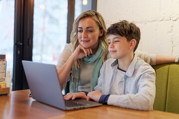 Mother and son sitting in a coffee shop, doing online studying or homework together on a laptop, mom helping her son