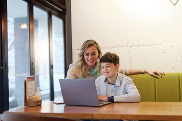 Mother and son sitting in a coffee shop, doing online studying or homework together on a laptop,...