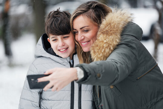 Young mom and her son taking a selfie on a snowy winter day outside in the park, having fun, sending pictures to their family and friends