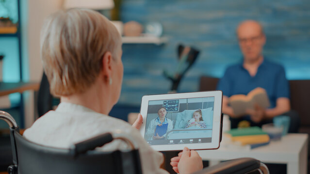 Elderly Person Holding Digital Tablet With Remote Videocall, Sitting In Wheelchair. Older Woman With Disability Talking To Doctor And Niece On Online Teleconference Chat, Using Device.