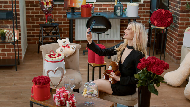 Blonde woman smiling and taking picture with teddy bear gift for valentines day celebration. Romantic surprise with flowers bouquet and sweet presents to celebrate love holiday.