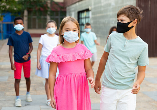 European boy and girl in face masks walking together hand in hand through city streets in summertime. Other kids walking behind.