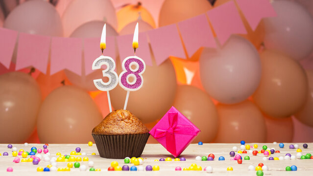 Decorations with balloons and a happy birthday candle with the number 38 for a woman. Happy birthday greetings in pink flowers for thirty-eight years for a girl, copy space. Muffin with a burning cand